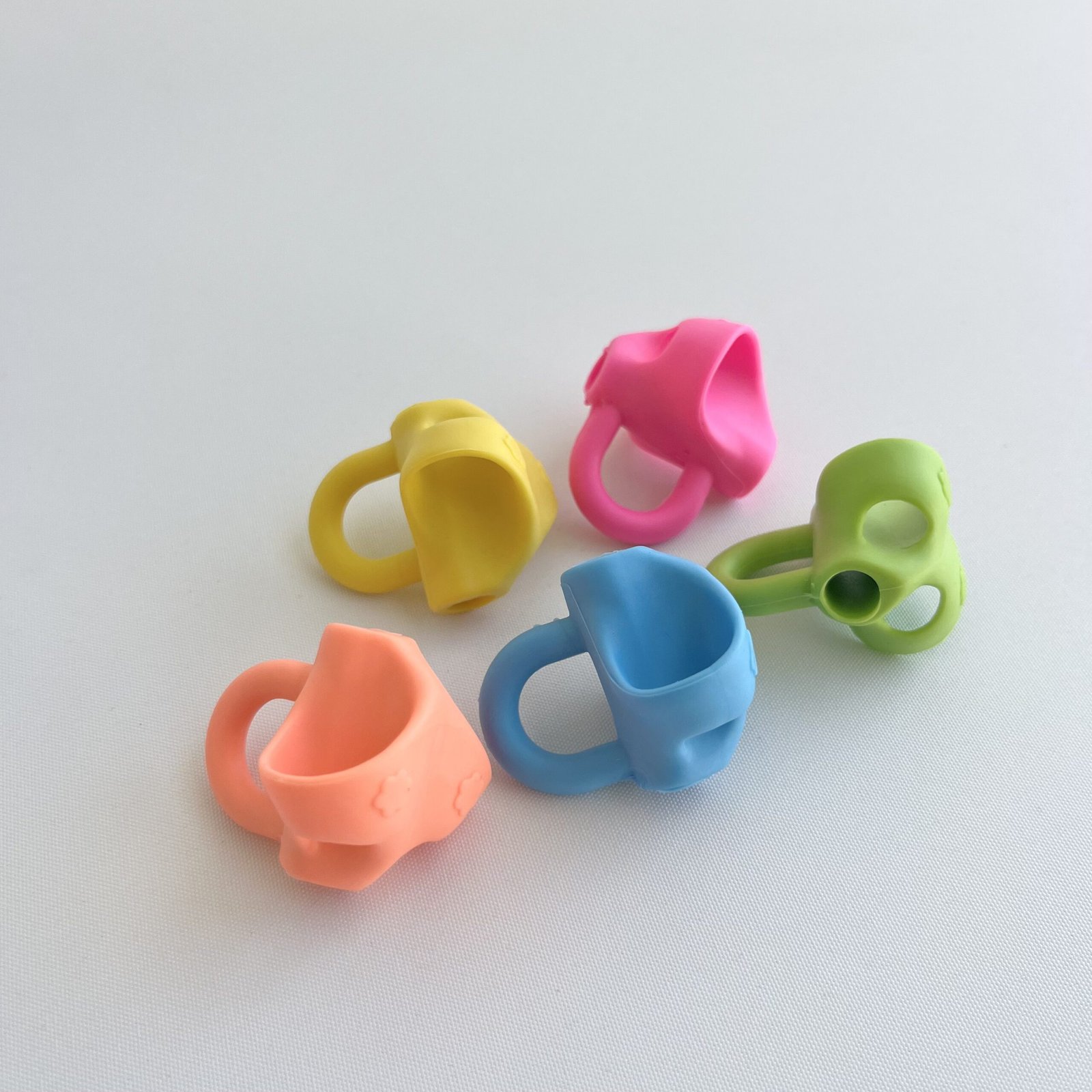 3-finger-pencil-grips Four colourful 3-finger pencil grips in yellow, pink, blue, and peach, scattered on a white background.
