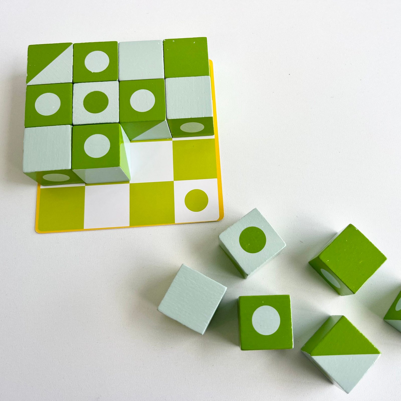 3D-jigsaw-puzzle-on-card A top-down view of a partially completed 3D Wooden Visual Perception Puzzle with green and white patterned cubes on a matching card. Several loose cubes are scattered beside it on a white background.