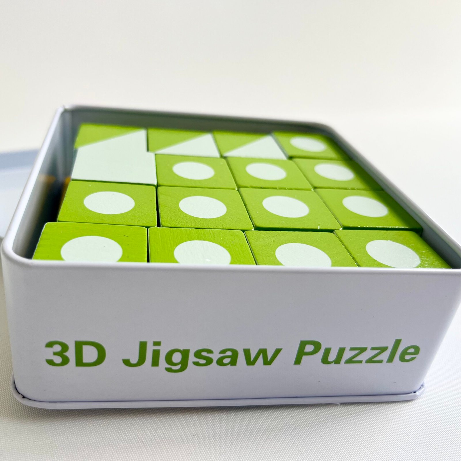 3D-jigsaw-puzzle A close-up, angled view of the 3D Wooden Visual Perception Puzzle set neatly packed inside a white metal tin, displaying green and white patterned wooden cubes. The tin has "3D Jigsaw Puzzle" written on its side.