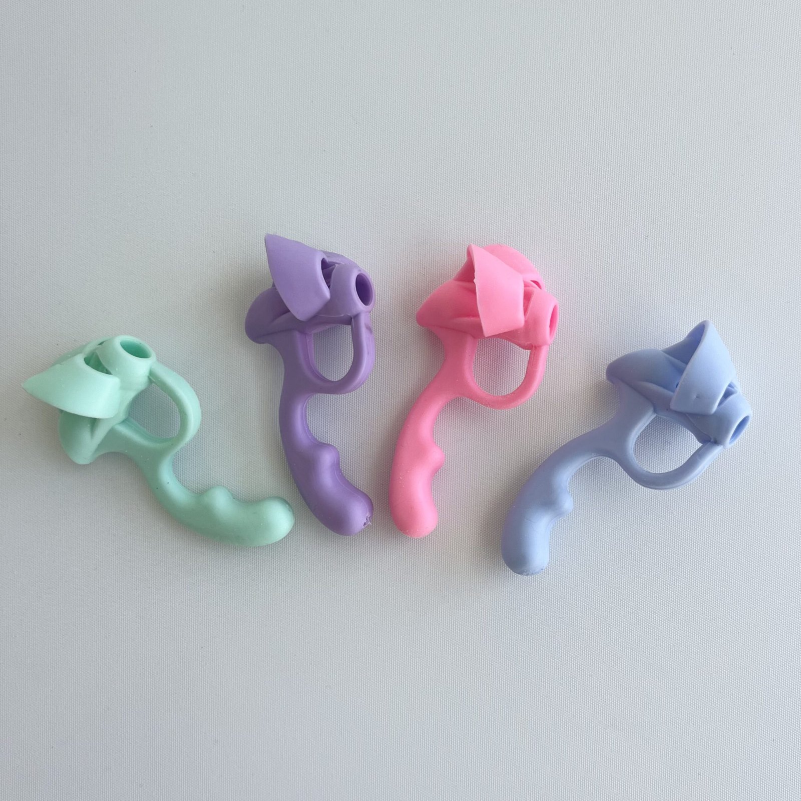 Four pastel-coloured 5-finger pencil grip correctors in a row on a white surface: mint green, lavender purple, pink, and light blue.