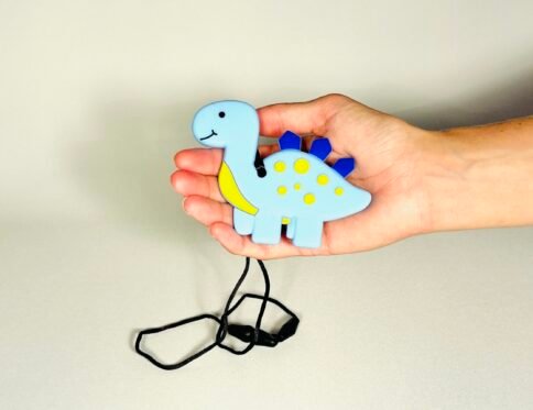 Child's hand holding a light blue and yellow dinosaur sensory chew necklace with a black safety cord.