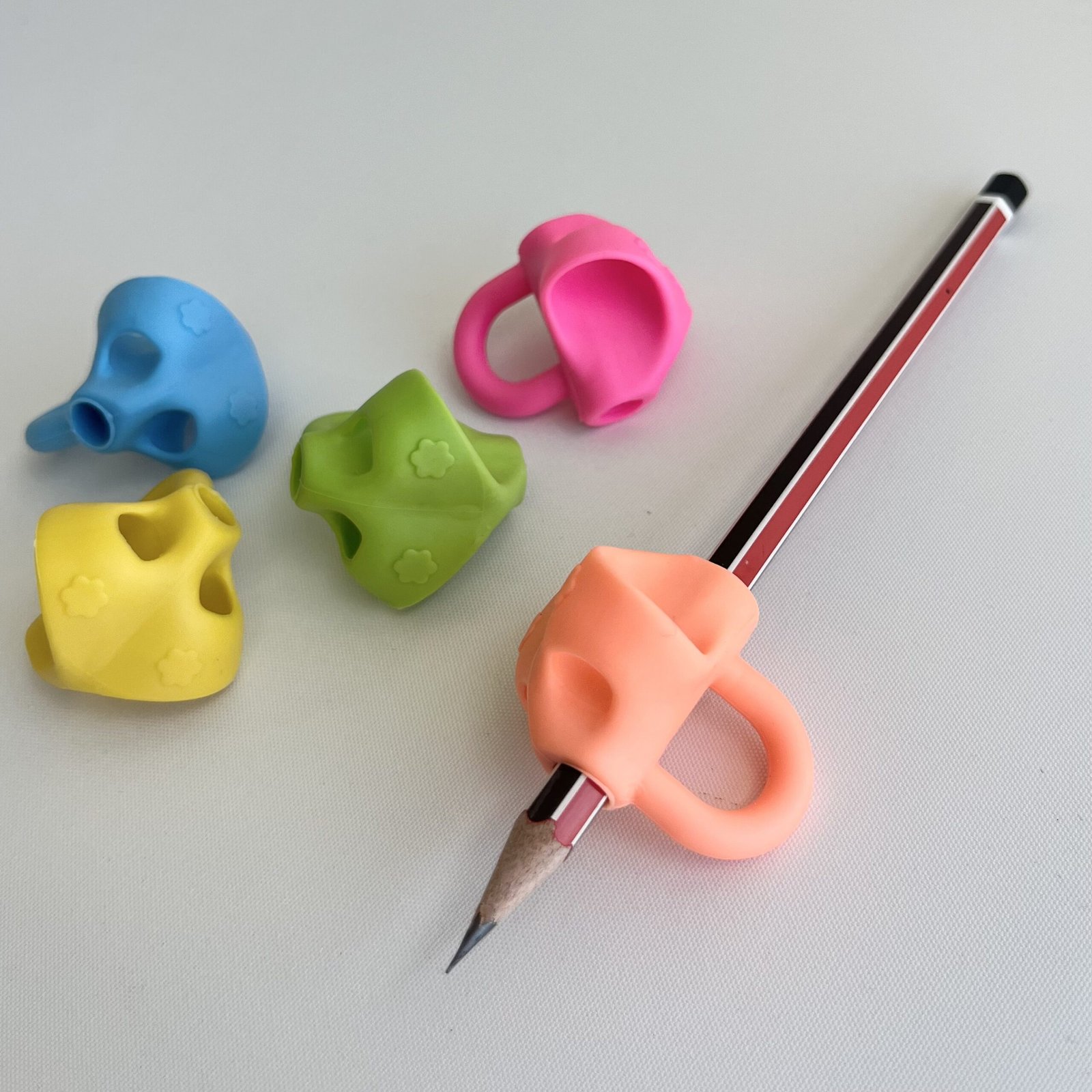 peach-3-finger-pencil-grip-on-pencil A sharpened pencil fitted with a peach 3-finger pencil grip, with three other colourful grips (blue, green, yellow) arranged behind it on a white surface.