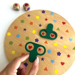 person-playing-wooden-shape-and-colour-matching-game A hand holding one green magnifier tool over two red heart shapes on the wooden I Spy game board. Another green magnifier tool is nearby, covering a red star and a red heart. Two wooden dice are in the background, showing a red heart and a blue star.