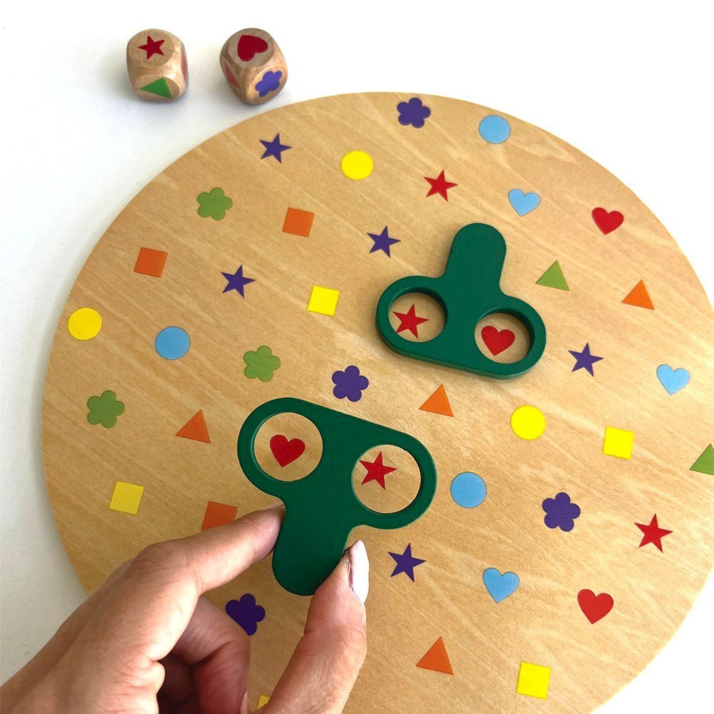 person-playing-wooden-shape-and-colour-matching-game A hand holding one green magnifier tool over two red heart shapes on the wooden I Spy game board. Another green magnifier tool is nearby, covering a red star and a red heart. Two wooden dice are in the background, showing a red heart and a blue star.