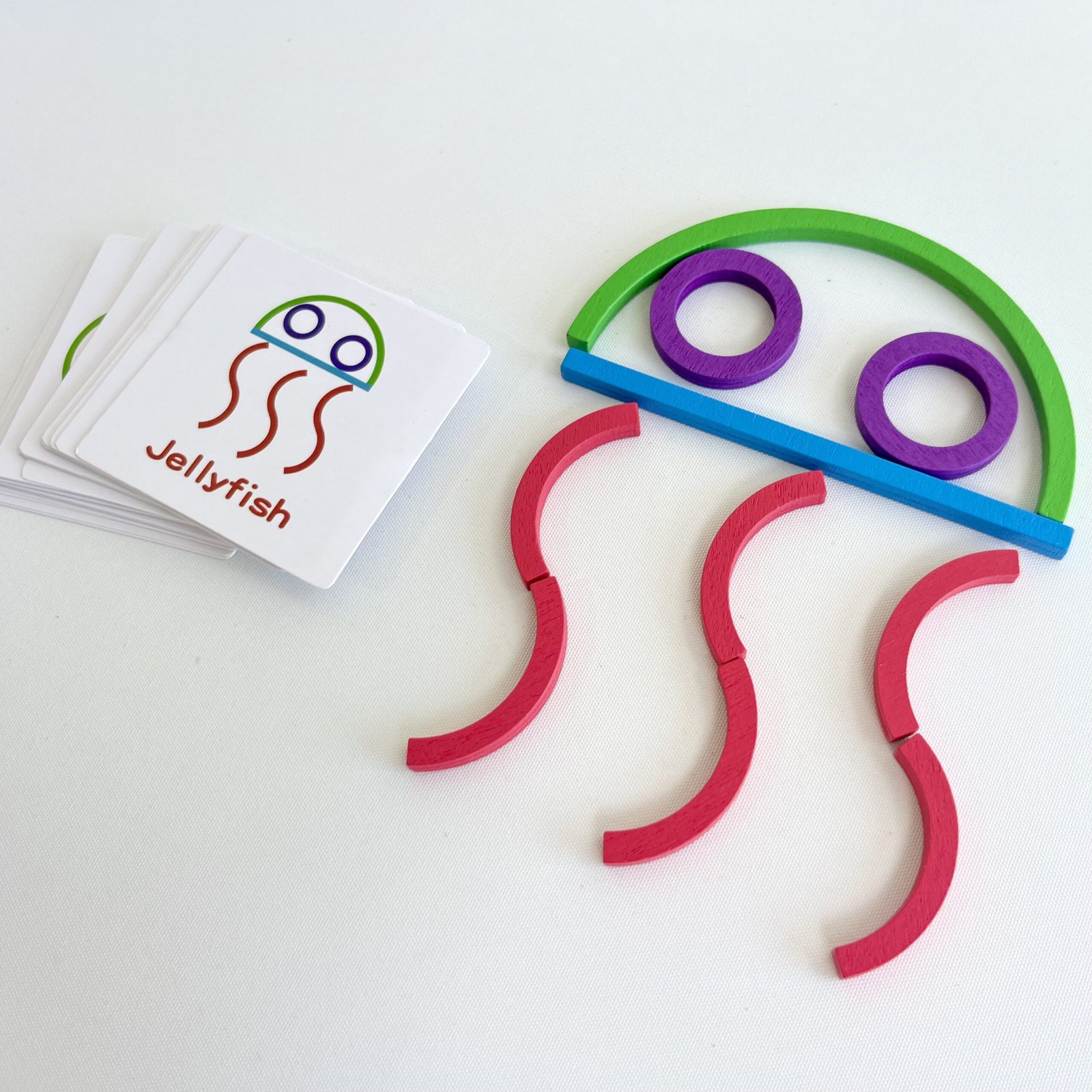 Top-down view of colourful wooden stick puzzle pieces arranged to form a jellyfish, with a stack of white challenge cards featuring a jellyfish image next to them.