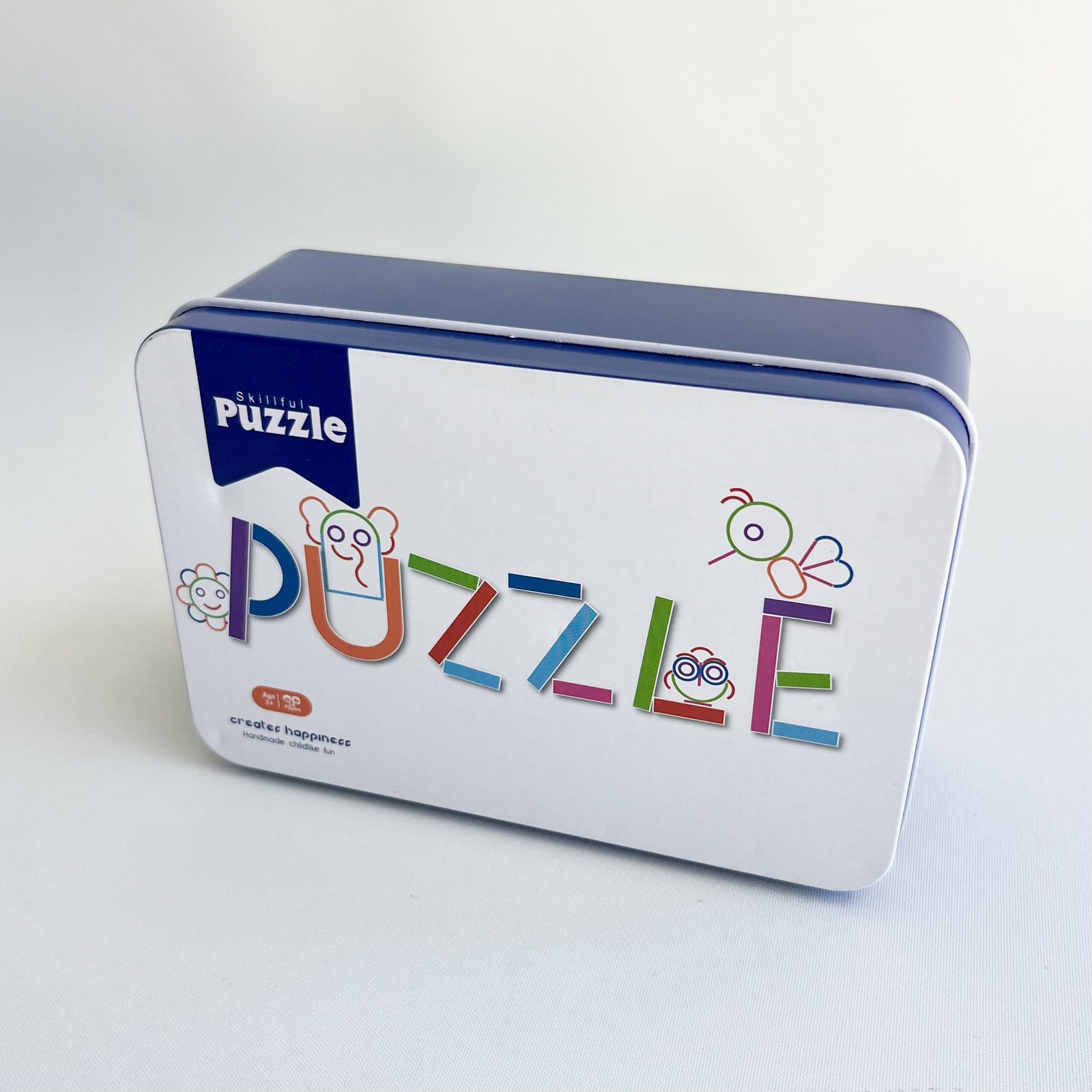 Close-up of the 'Skilful Puzzle' tin, a blue-lidded, white metal box with colourful 'Puzzles' lettering and small cartoon figures on the front.
