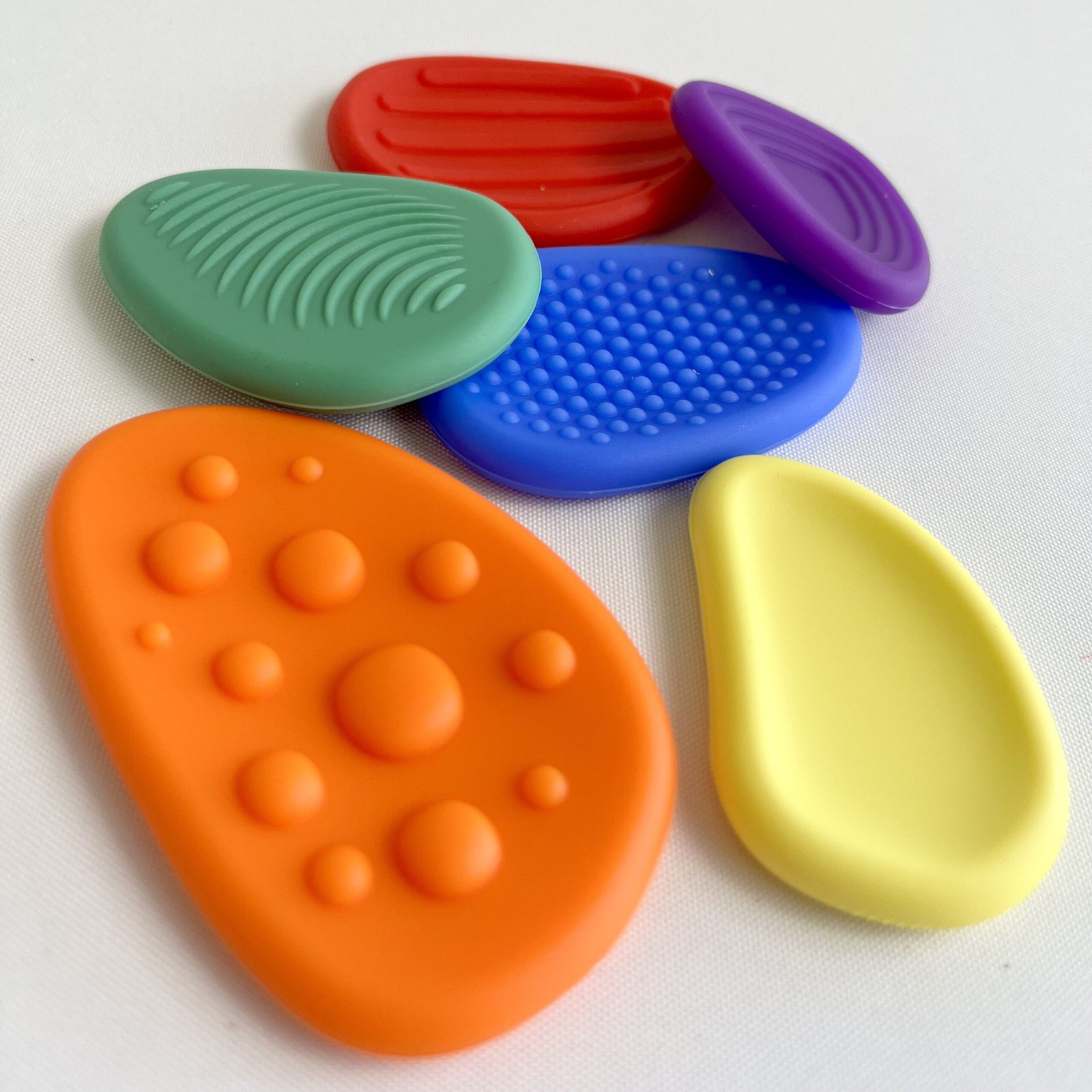 textured-fidget-stones Angled close-up of six textured silicone fidget stones scattered on a white background, showing their unique tactile patterns and vibrant colours (red, blue, green, orange, yellow, purple). Ideal for sensory exploration.