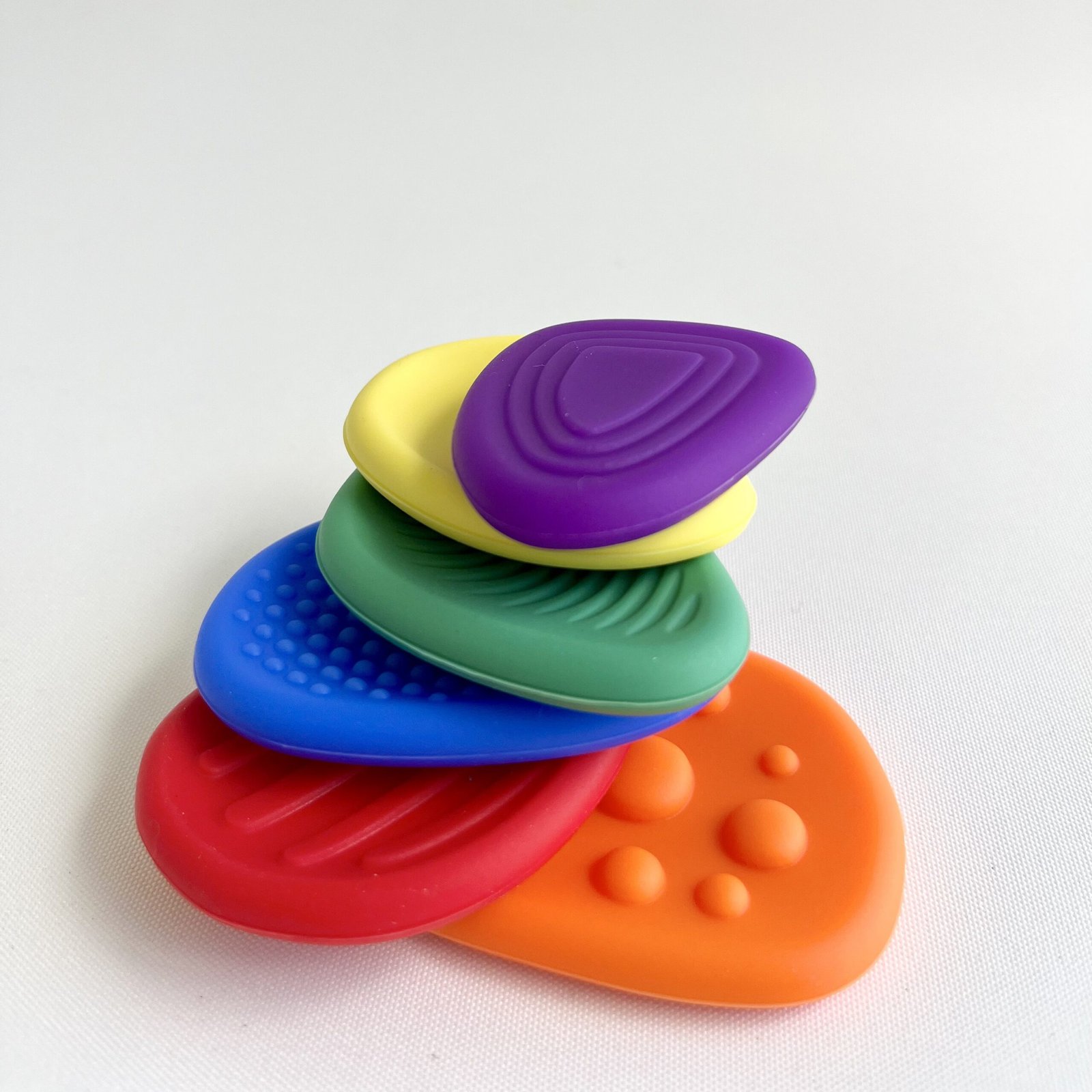 textured-fidget-stones-stacked Stack of five textured silicone fidget stones, from bottom to top: orange, red, blue, green, yellow, with a purple stone resting on top, all displaying their unique patterns against a white background. These fidgets promote focus.