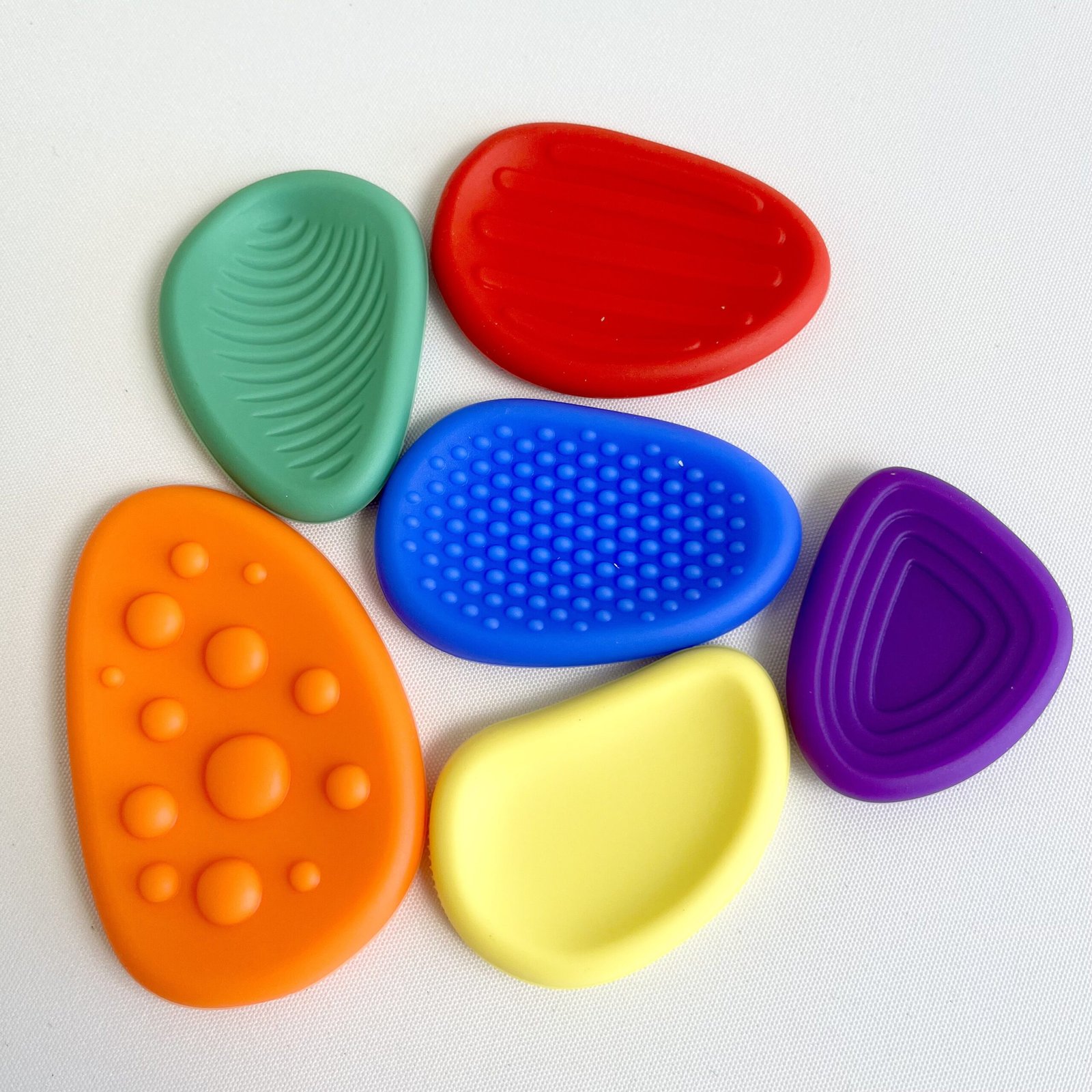 textured-fidget-stones-variety Top-down view of six textured silicone fidget stones in red, blue, green, orange, yellow, and purple, arranged in a flower shape on a white background. Each stone has a unique tactile pattern for sensory input.