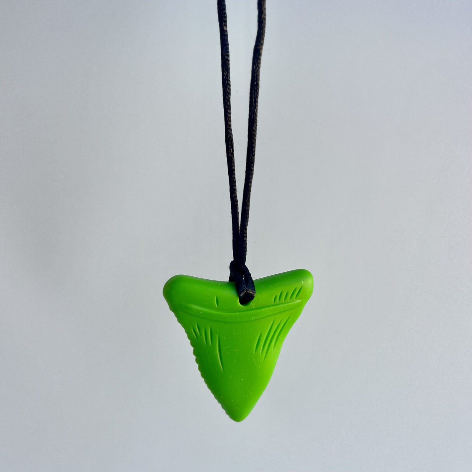 A single green chewable necklace shaped like a shark tooth, hanging on a black cord against a light grey background.