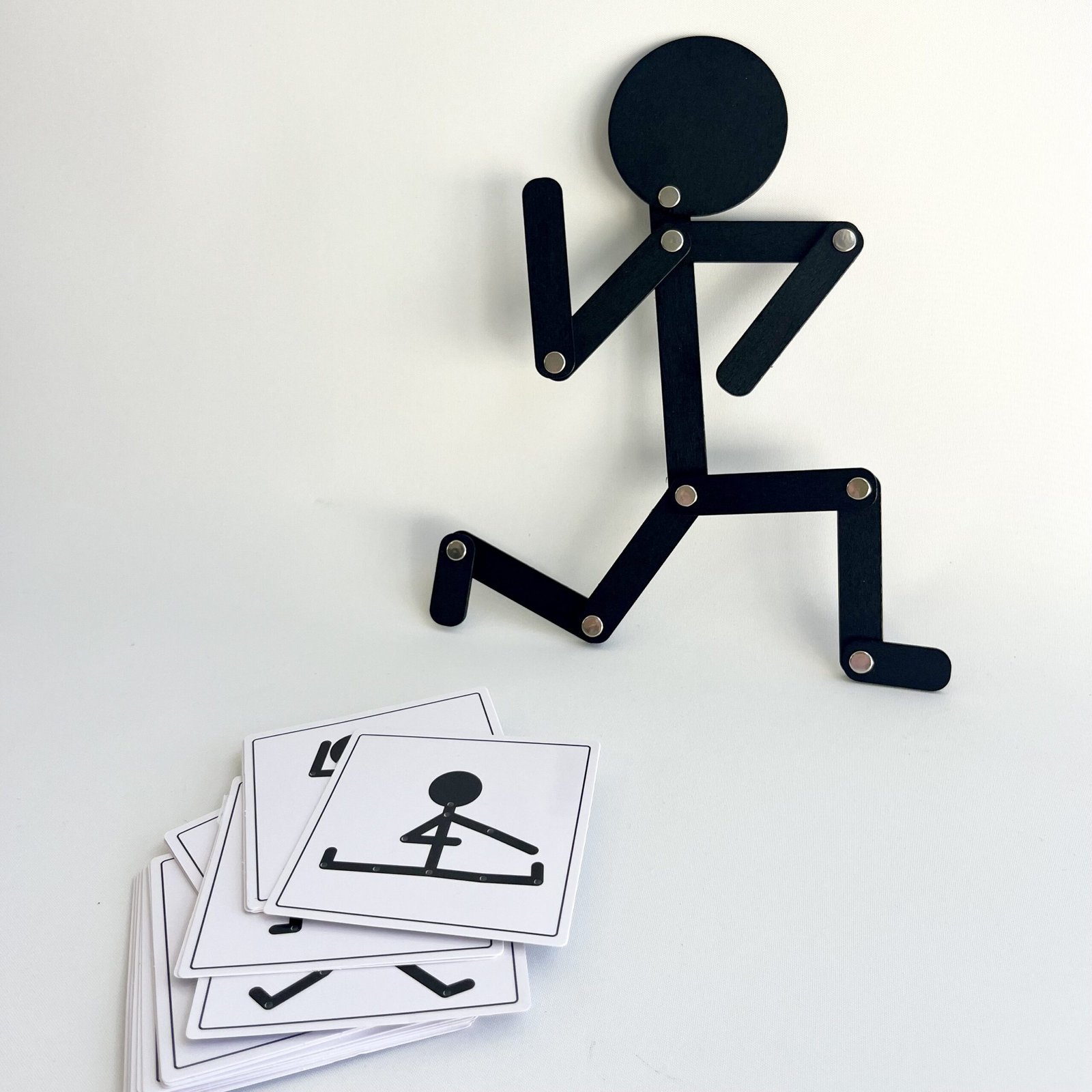wooden-man-running Overhead view of the black Pose & Play Wooden Man articulated figure in a running pose, with a stack of black and white body pose challenge cards lying below it on a white background.