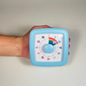 A child's hand holding a light blue square visual countdown timer with a white face, black numbers, and a central light blue knob, displaying a rainbow segment for 10 minutes.