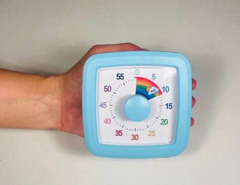 A child's hand holding a light blue square visual countdown timer with a white face, black numbers, and a central light blue knob, displaying a rainbow segment for 10 minutes.