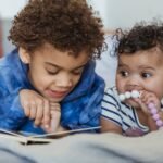 Cute African American boy reading book while lying on sofa with little sibling in light room at home on blurred background