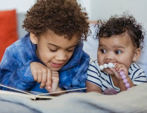 Cute African American boy reading book while lying on sofa with little sibling in light room at home on blurred background