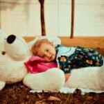 A cute toddler in a floral dress hugging a large stuffed teddy bear on grass.