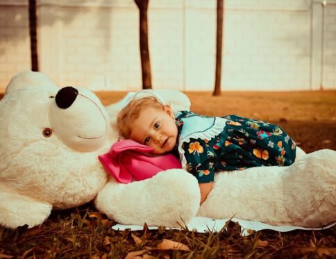 A cute toddler in a floral dress hugging a large stuffed teddy bear on grass.